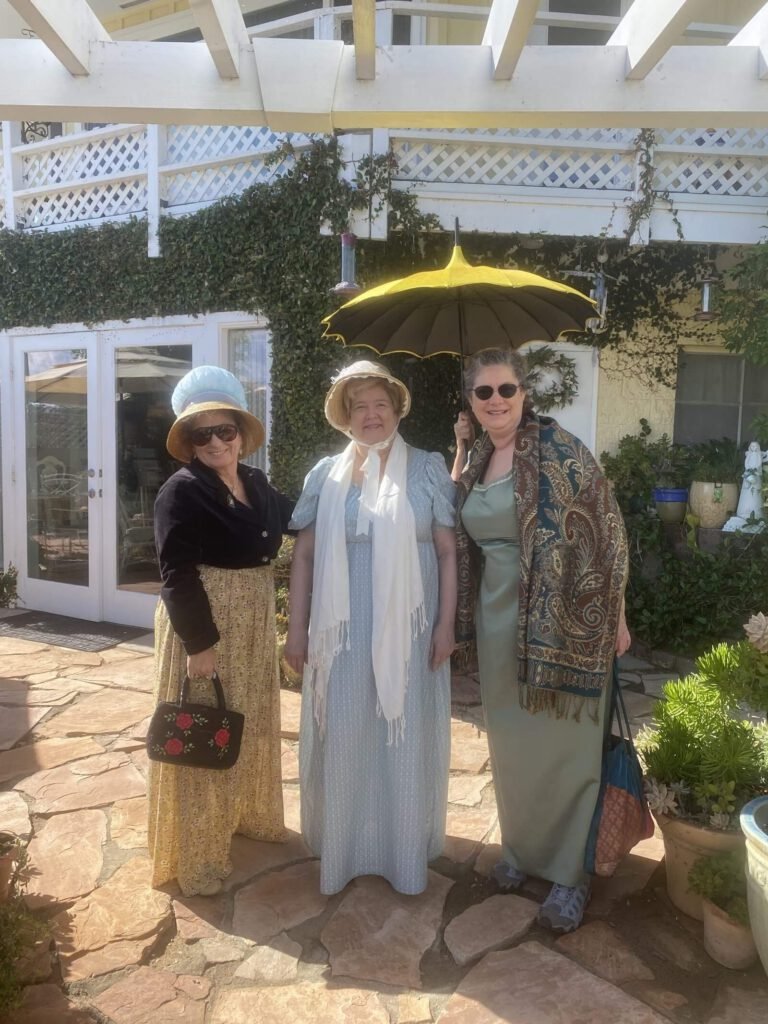 Three women dressed in very elegant clothes, wearing scarves and hats, and carrying a very fancy umbrella, standing in front of Claudia's tea house.