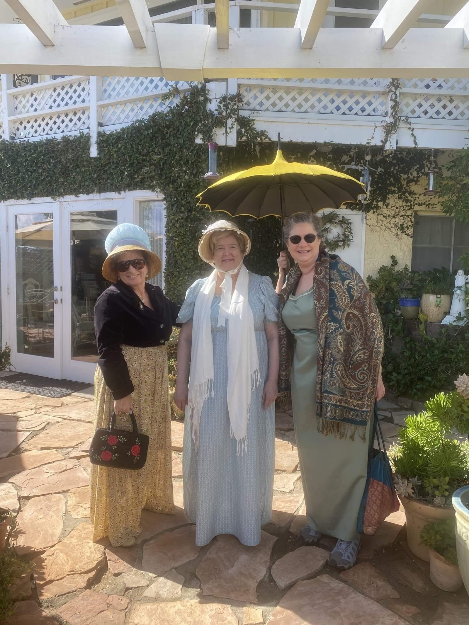 Three women dressed in very elegant clothes, wearing scarves and hats, and carrying a very fancy umbrella, standing in front of Claudia's tea house.